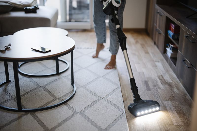 A person uses a vacuum cleaner on the floor in a living room. The scene shows furniture such as a table and a phone nearby. Bright light comes through a window.