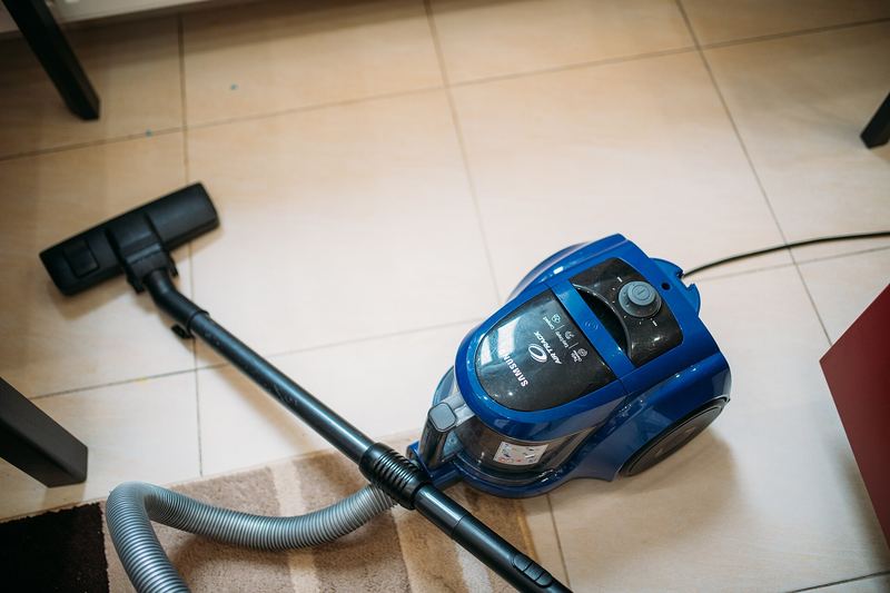A vacuum cleaner sits on the floor near a rug in a living room. The room is tidy and organized, showing the process of cleaning. Light enters through a window, 